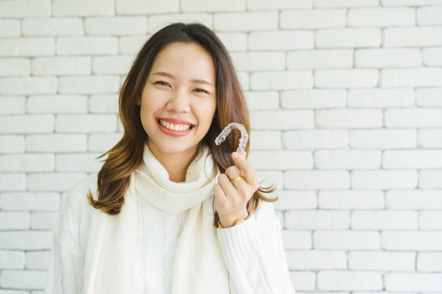 Brunette woman in a turtleneck sweater smiles while holding her Invisalign clear aligners