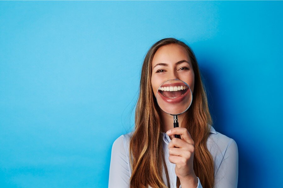 Brunette woman holds a magnifying glass to her teeth while standing against a blue wall
