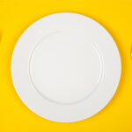 Aerial view of a white plate flanked by silverware on a yellow background