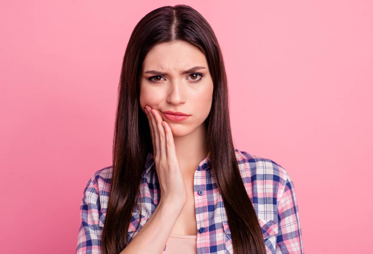 A brunette woman in a plaid shirt cringes in pain and touches her cheek against a pink wall