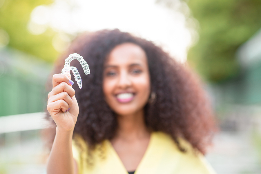 Blurred Black woman in a yellow blouse smiles as she holds up her Invisalign aligners